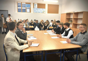 Uczniowie siedzą przy stole podczas pokazowej debaty oksfordzkiej w tle widownia